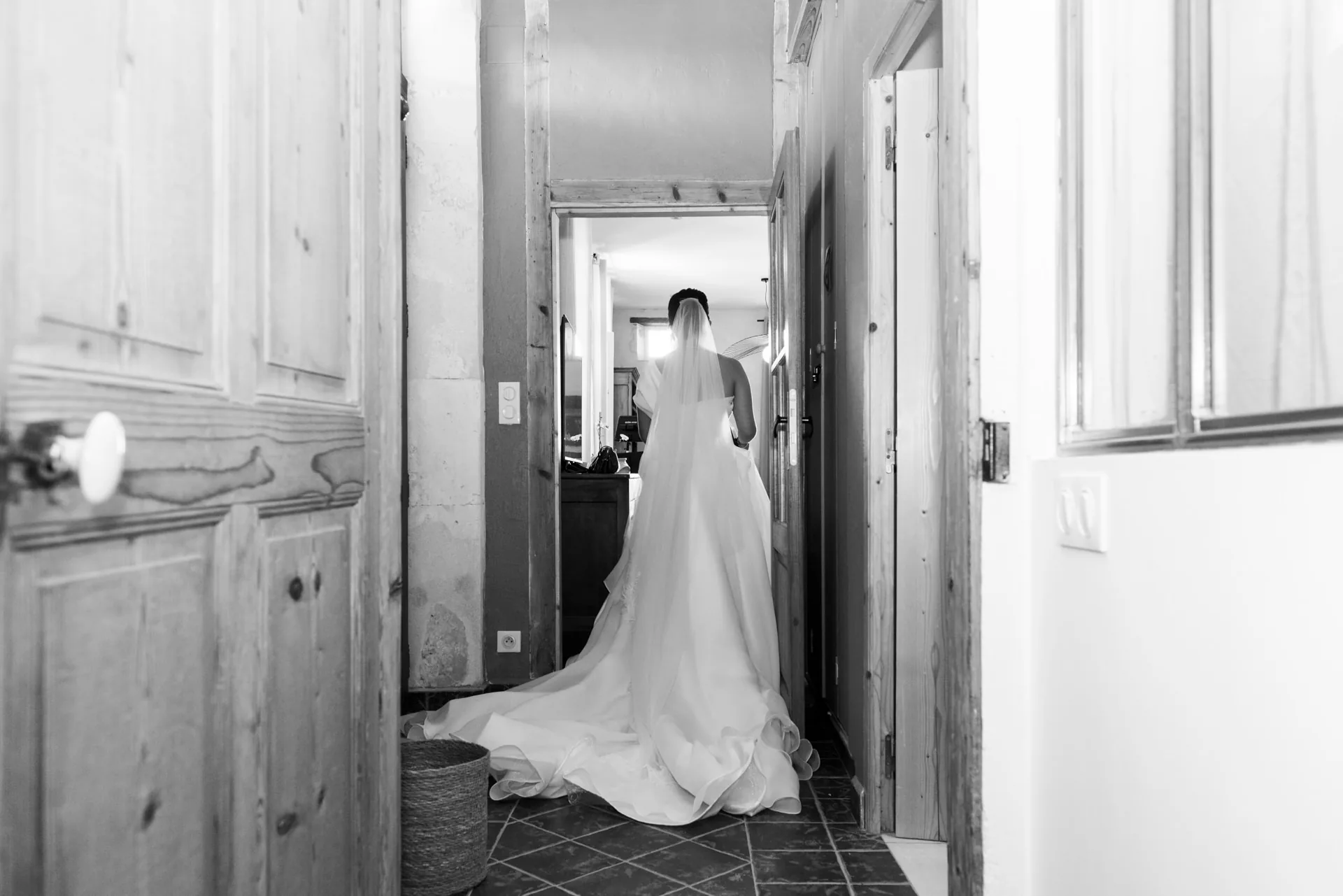 La mariée de dos traverse un couloir en pierre, sa traîne déployée derrière elle, en noir et blanc