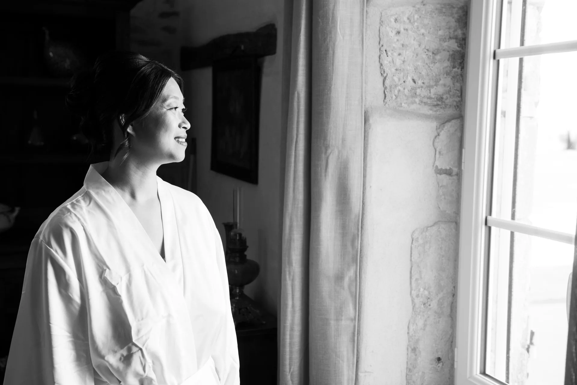 La mariée en peignoir blanc regarde par la fenêtre, en noir et blanc