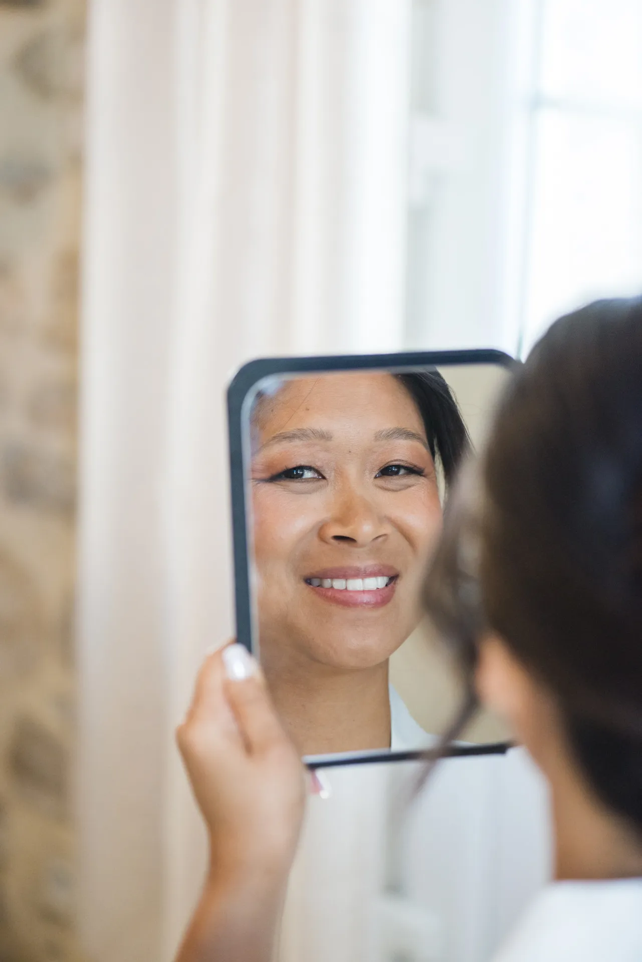 La mariée sourit en se regardant dans un petit miroir après son maquillage