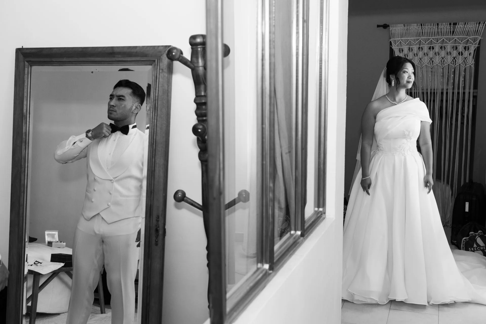 Le marié ajuste son nœud papillon tandis que la mariée apparaît dans l’encadrement d’une porte