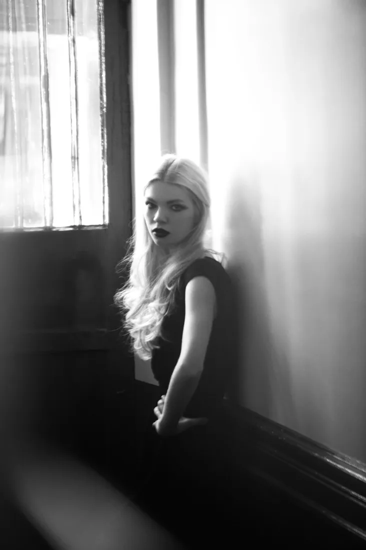 Modèle blonde photographiée en noir et blanc près d’une fenêtre